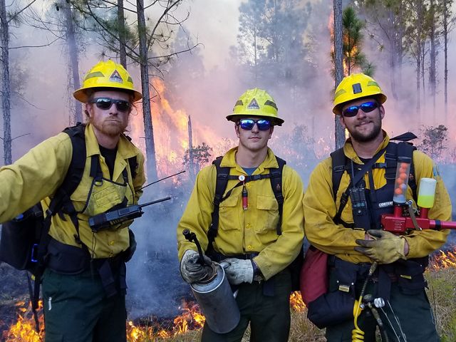 Three students from the University of Idaho dressed in fire-protective gear pose near a prescribed fire at Tiger Creek Preserve.