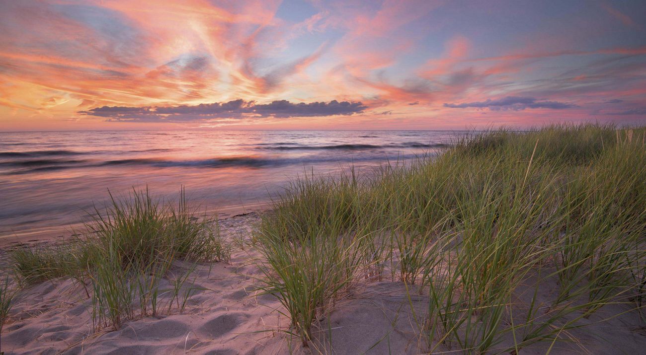 A serene sunset over a beach with vibrant sky colors reflected on the lake surface, foregrounded by sand dunes with tall grasses.