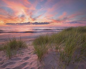 A serene sunset over a beach with vibrant sky colors reflected on the lake surface, foregrounded by sand dunes with tall grasses.