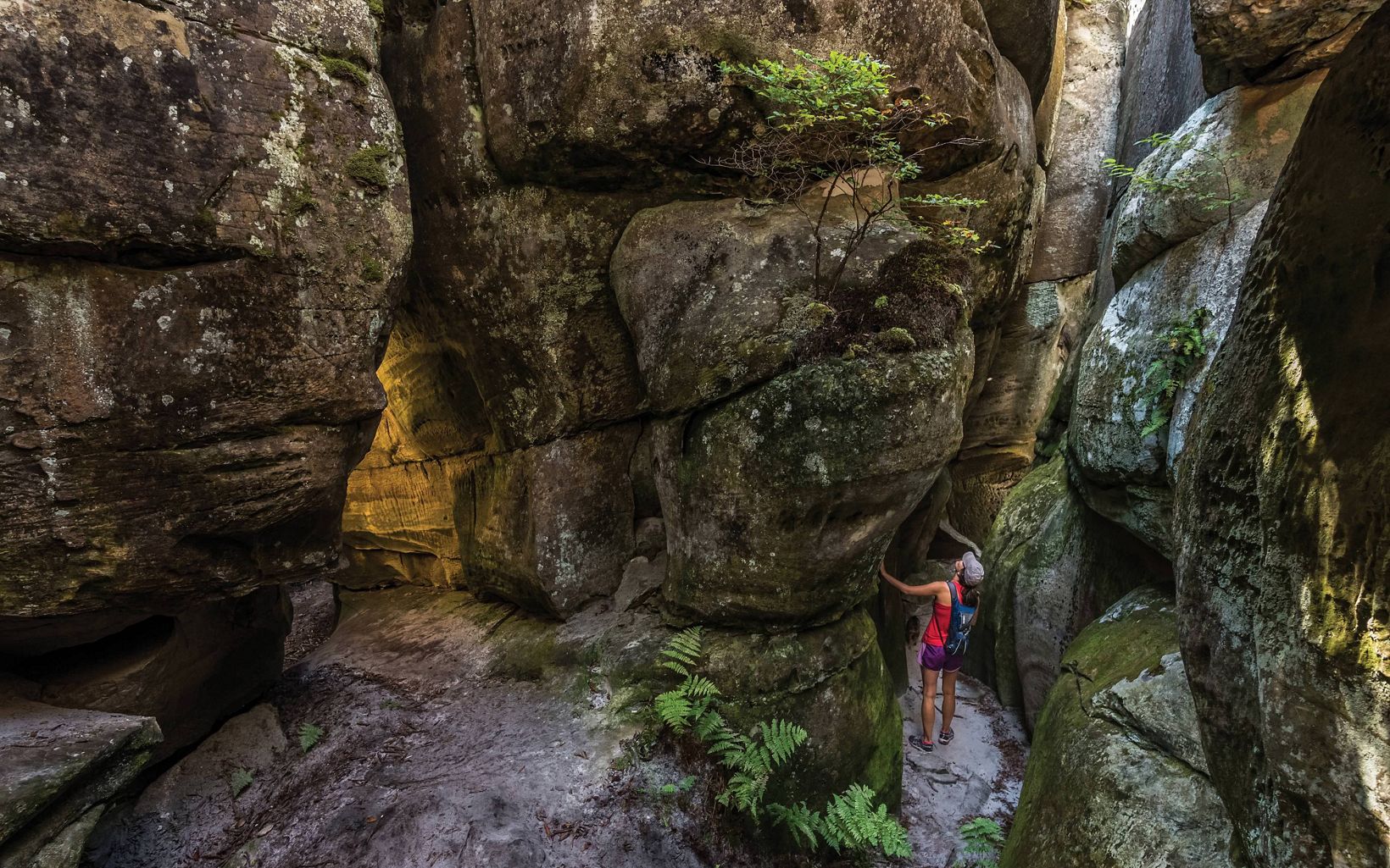 A woman in a pink shirt rests her hand on the face of a large rock formation. She looks up from the narrow slot created by the boulders.