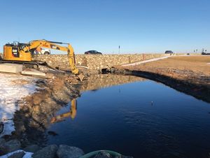 A backhoe clears out around a small stone box culvert in a pond.