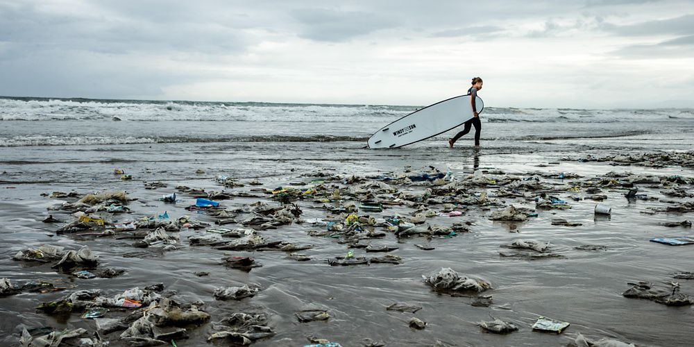 A surfer walks along a trash-filled beach.