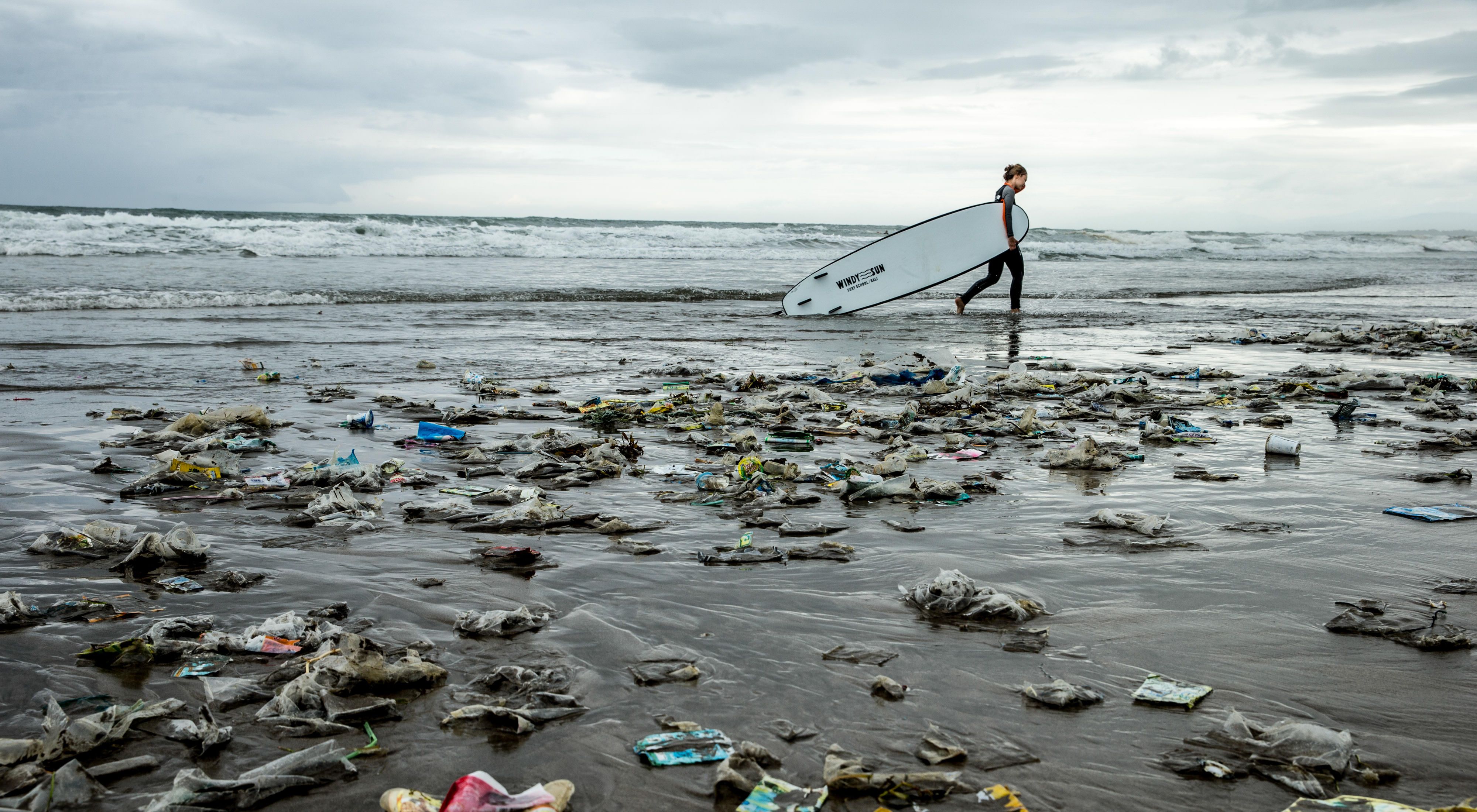A surfer walks along a trash strewn beach. 