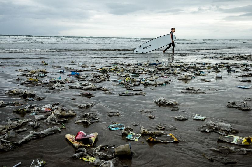 A person carrying a surfboard walks along a beach that is strewn with trash and plastic.