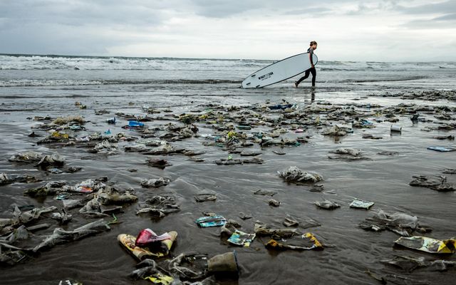 A surfer walks on a beach covered in trash.