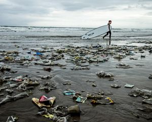 Surfer walking past garbage strewn on a beach.