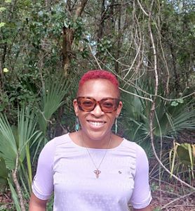 A woman with short red hair and sunglasses stands in front of palm fronds.