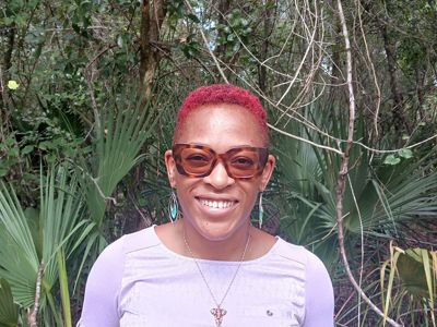 A woman with short red hair and sunglasses stands in front of palm fronds.