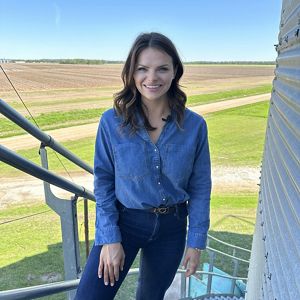 An agricultural field is the backdrop for a woman smiling for a photograph. 
