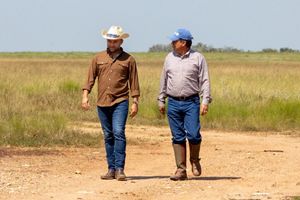 Two men walk along a dirt path in a field of green grass.