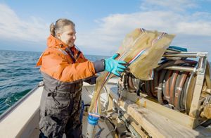 A middle-aged woman in an orange and black jumpsuit handles bag-shaped scientific equipment at the back of a research vessel on the ocean
