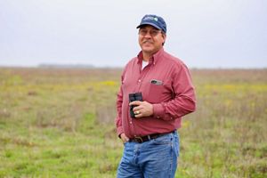 A man wearing a hat stands in a field of green grass, holding binoculars at his side.
