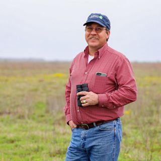 A man wearing a hat stands in a field of green grass, holding binoculars at his side.