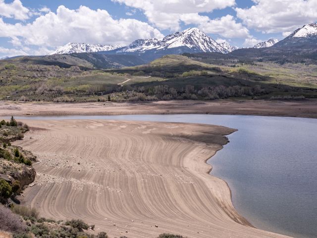 Low water levels in a reservoir expose lines of sediment., with snow-capped mountains in the distance.