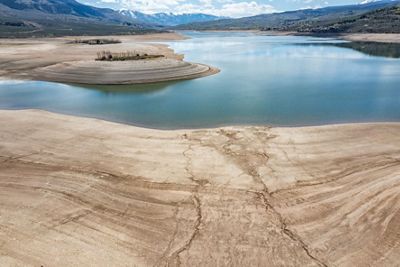 View of a reservoir with a low amount of water that is reflecting the blue sky, with beige rock formations around it and snow-capped mountains in the background.