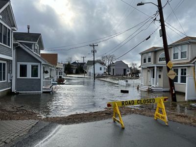 A coastal road in Hull with typical multi-unit homes on either side of the street, covered in several inches of water from king tide flooding. A "Road Closed" sign blocks the street.