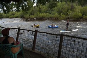 Two people in kayaks and one person on a stand-up paddleboard travel down a river as people on a balcony look on.