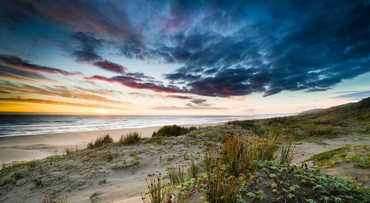The sun sets over sand dunes along a beach.