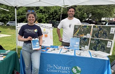 Emma Gildesgame and Andrew Laquerre stand at a table with a TNC-branded tablecloth and materials, as well as flood maps of Taunton, under a tent on the grass at a festival.