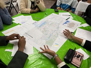 Many pairs of hands hover over maps sitting on a table.