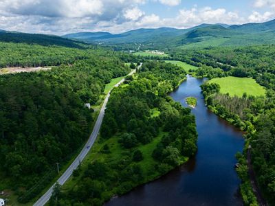 A blue river splits the landscape of lush green forests and a single road on each side with a silhouetted mountain range in the distance below a sky with fluffy clouds.