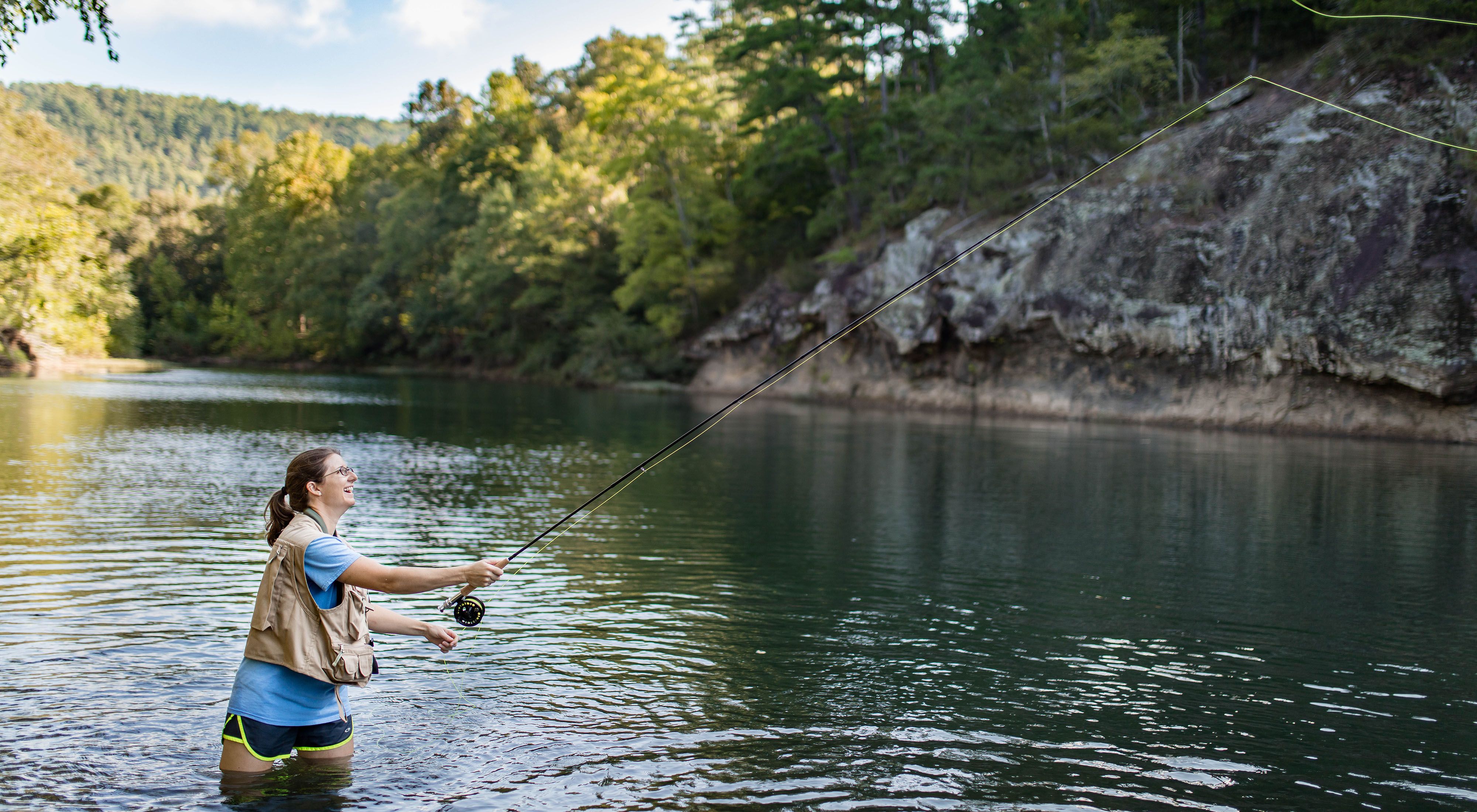 A smiling woman in a fishing vest and waders casting a line on the edge of a river, with rock bluffs in the background.