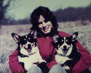 A person holds two corgi's under each of their arms and smiles at a camera. 