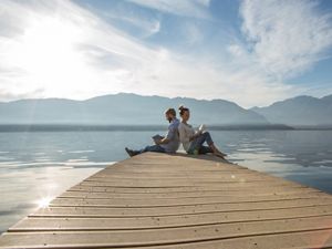 A man and a woman sit back to back at the end of a pier. They're each reading a book. The water behind them stretches to the horizon where it meets a mountain ridge.