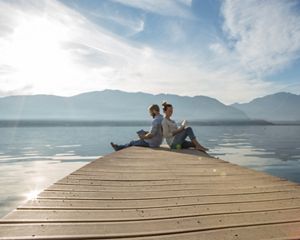 A man and a woman sit back to back at the end of a pier. They're each reading a book. The water behind them stretches to the horizon where it meets a mountain ridge.