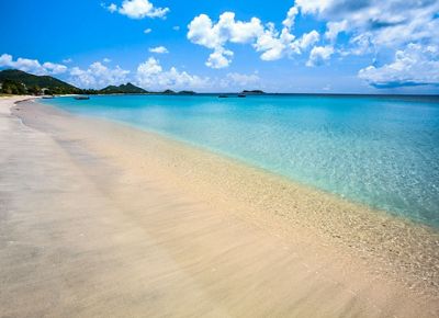 A sandy beach leads to deep blue water, with green mountains on the horizon and boats dotting the shore in the background.