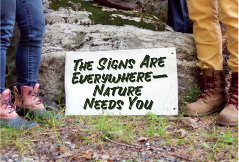 Two people wearing hiking boots frame a sign that encourages involvement in nature conservation.