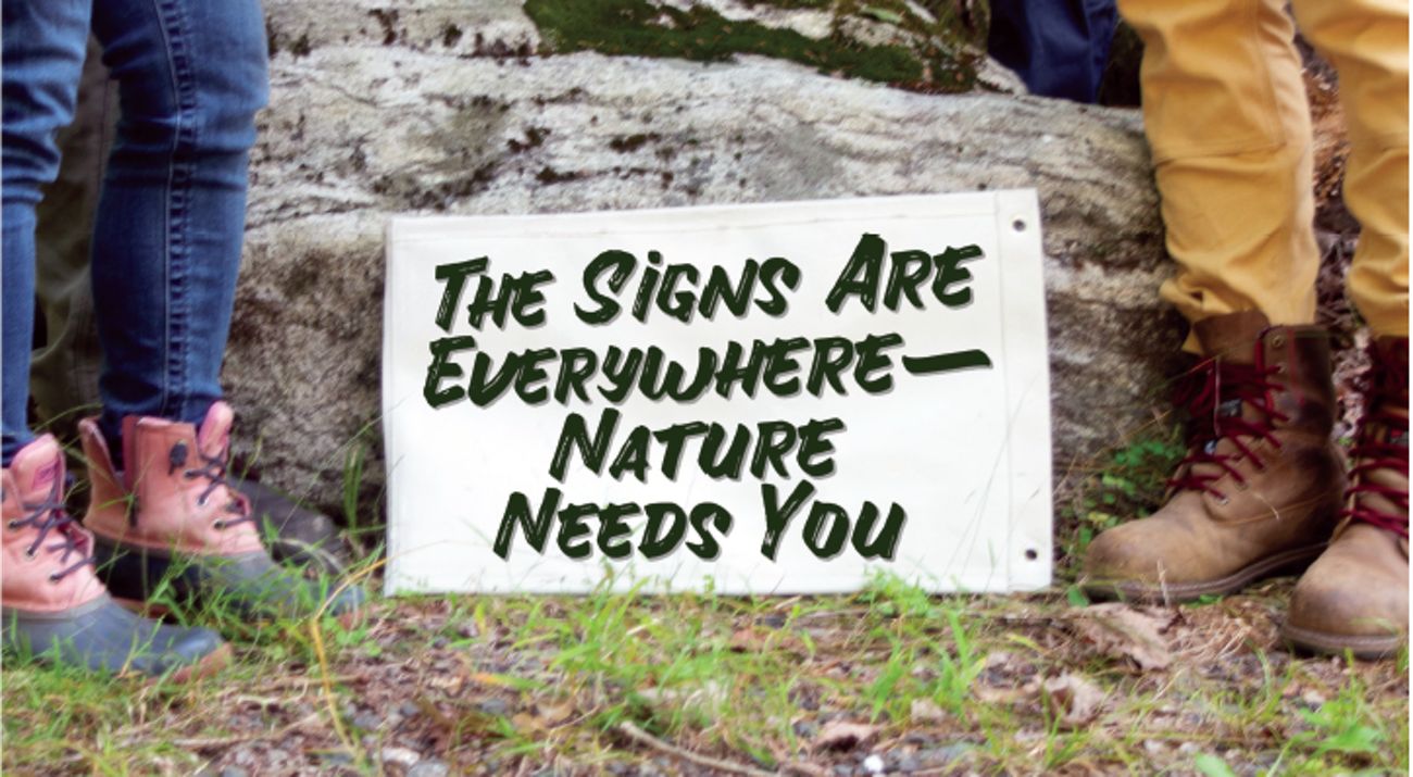 A sign is situated between the legs of two people wearing outdoor footwear.