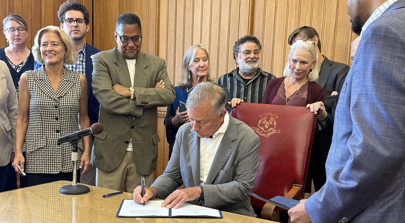 A group of people surround a man sitting at a desk signing a document.