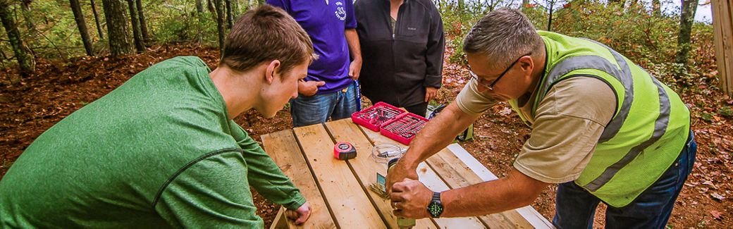 Four people bend over a picnic table holding tools.