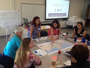 Several women gather around an indoor table and examine a map on the table.