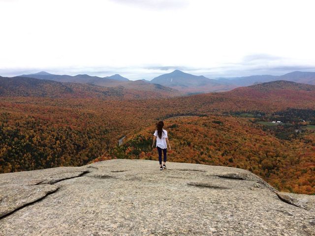 A person standing with back to camera on a rock facing the Adirondacks.