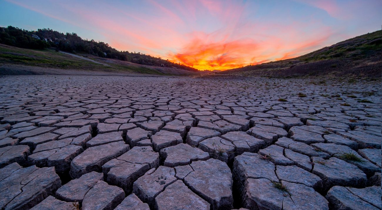Lecho de río seco y agrietado con el atardecer de diferentes colores en el fondo.