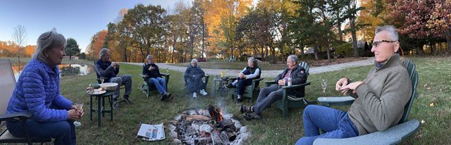 A group of people sits in a circle around a small outdoor fire near a pond, with colorful autumn trees in the background.
