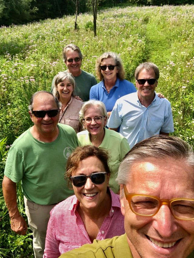 A group of people stands together in a sunny meadow filled with wildflowers, surrounded by lush green landscape.