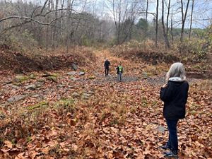 People walk along a rocky creek bed covered with fallen leaves in a quiet forest during late autumn.