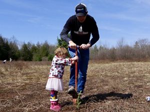 A man and toddler girl in a pink tutu use a long handled digging tool to plant a tree seedling. They are in an open filled ringed by tall cedar trees.
