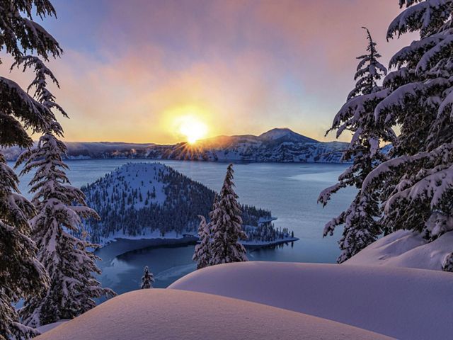 The sun rises in the morning over snowy Crater Lake National Park in Oregon. There's a view of the lake and Wizard Island.