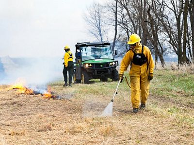 A burn crew worker spraying water along a fireline with a vehicle and another crew member in the background.