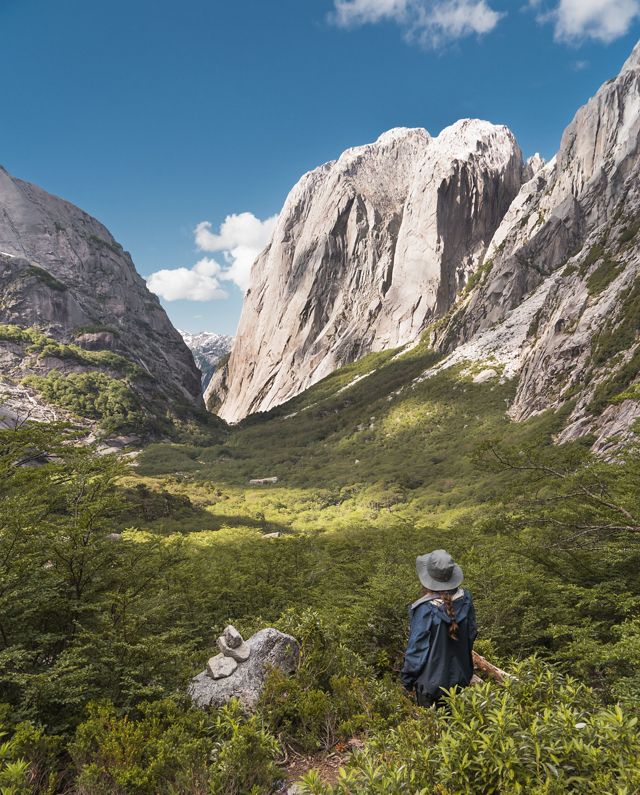 A person stands and looks out toward a wide green valley flanked by towering mountains.
