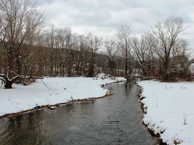 A wide creek flows between snow covered banks. A mountain ridge is visible in the background behind a row of tall leafless trees. Heavy clouds hang low in the sky.
