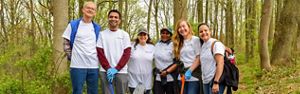 Six people pose together during a stream cleanup event. They are holding large black plastic trash bags and carrying long orange grabbers.
