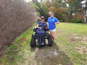 A person in a wheelchair with tank-like treads is smiling and wearing a red cap and sunglasses, holding a leash for a dog in front. Another person stands beside, also smiling, wearing a blue shirt and sunglasses. They are outdoors on a grassy path with trees and bushes around.