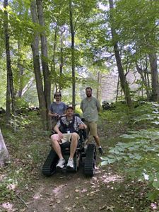 Three individuals are on a wooded trail with trees and greenery. One person sits in a rugged wheelchair designed for off-road use. The other two stand nearby on the path, surrounded by dense foliage.
