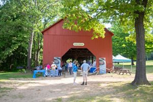 A rustic red barn labeled "Picnic Shelter" is surrounded by tall trees. Nearby, people are engaged in various activities, and a white and green canopy tent is set up. A few picnic tables are visible.