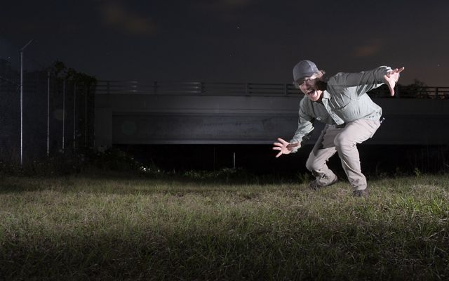 At night, man kneels and raises arms at camera, in front of overpass